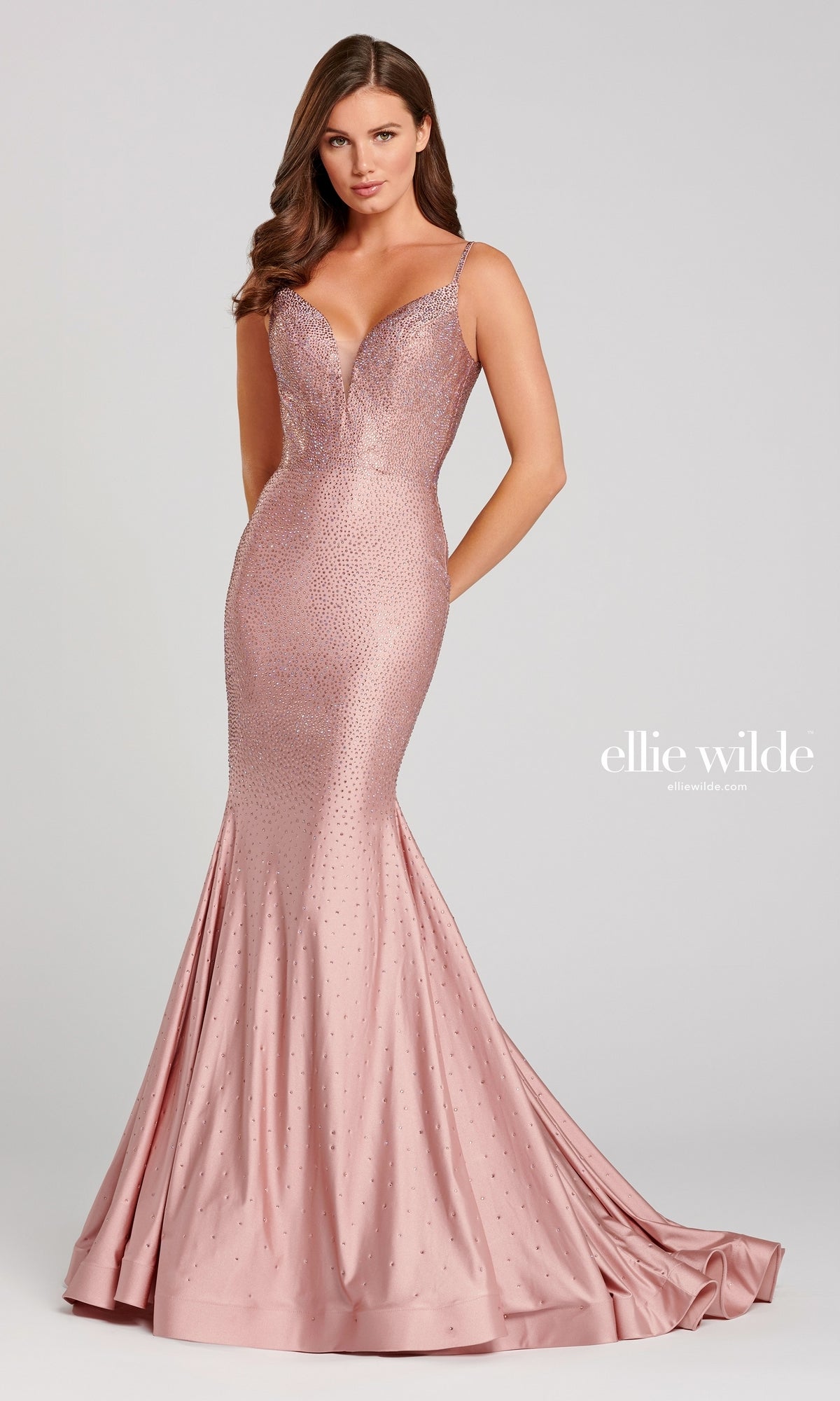 A woman wears the Ellie Wilde Beaded Mermaid Prom Dress with Cut Out—a fitted, blush pink gown featuring a v-neck, thin straps, and subtle embellishments—standing against a plain background.
